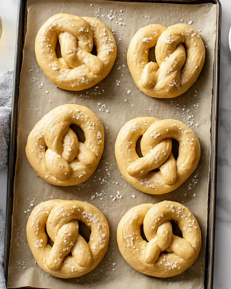 Several freshly unbaked gluten-free Bavarian soft pretzels with coarse salt on a parchment-lined tray.