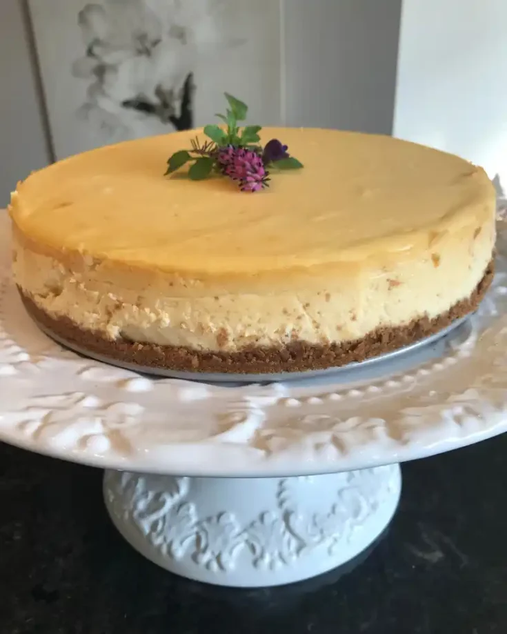 A rich and creamy gluten-free New York cheesecake with a golden-brown crust, topped with fresh berries and a drizzle of caramel, served on a white plate.