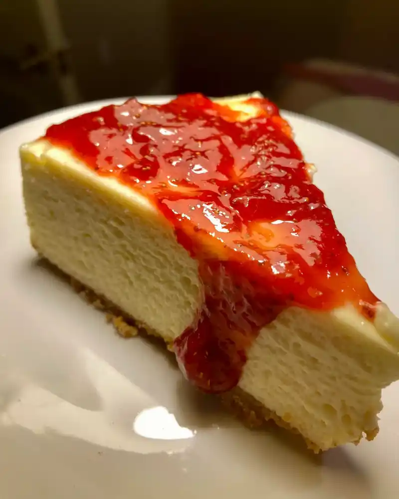 A rich and creamy gluten-free New York cheesecake with a golden-brown crust, topped with fresh berries and a drizzle of caramel, served on a white plate.