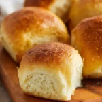 Soft, golden gluten-free dinner rolls on a rustic wooden board, brushed with melted butter and showing a fluffy, tender crumb inside.