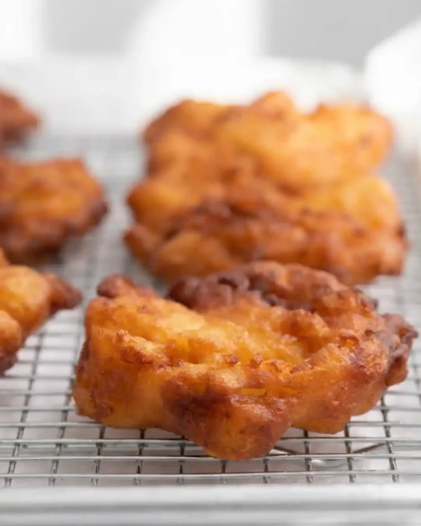 Gluten-free apple fritters cooling on a wire rack after frying, with golden brown, craggy edges and a soft, tender center.