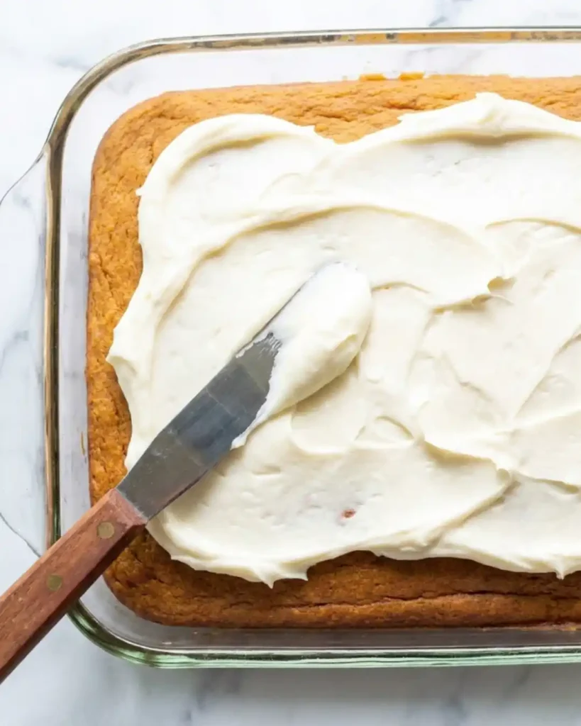Cream cheese frosting spread with an offset spatula over gluten-free pumpkin cake in a glass 9x13 pan